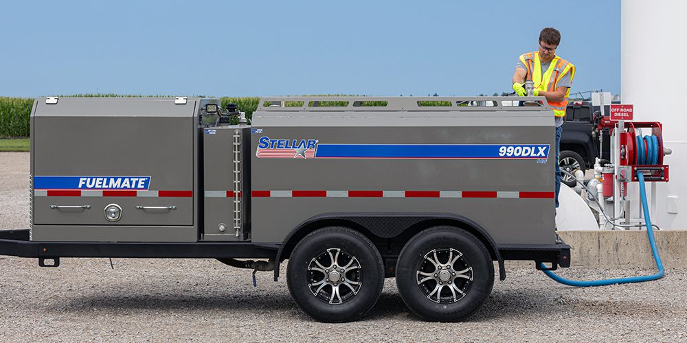 A person wearing a yellow and orange high-visibility vest is filling a Stellar FuelMate 990DLX Deluxe Fuel Trailer. In the background, there is a field of corn.