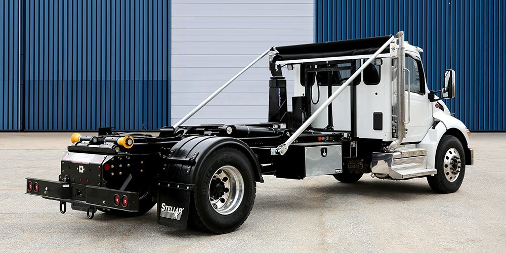 Stellar NXT24 hooklift mounted on a white truck chassis, parked in front of a blue industrial building.