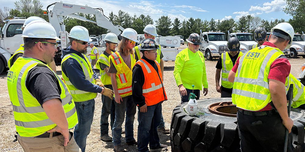 A group of service technicians in high-visibility vests and hard hats gathering around a large OTR tire during a Tire Industry Association technical training session in front of a Stellar Industries crane truck.