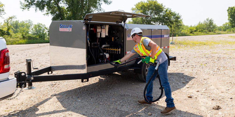 Operator pulling fuel hose from an open compartment of a grey Stellar Multi-Tank FuelMate® trailer at a jobsite.