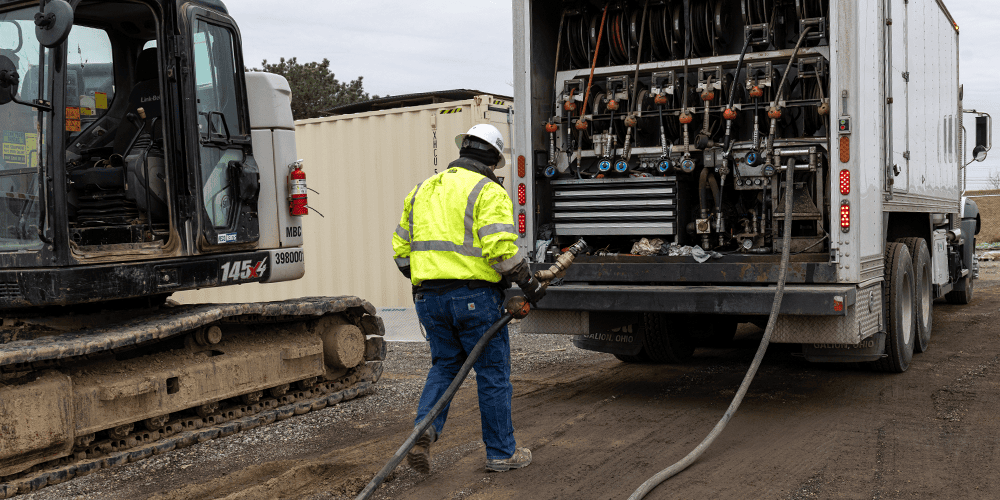 Technician pulling a service hose from the rear open hatch of a white Stellar Fuel & Lube Truck toward an excavator.