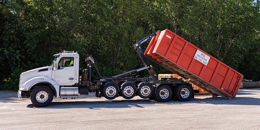 A white heavy-duty truck using a Stellar Hooklift System to tilt and unload a large red roll-off container.