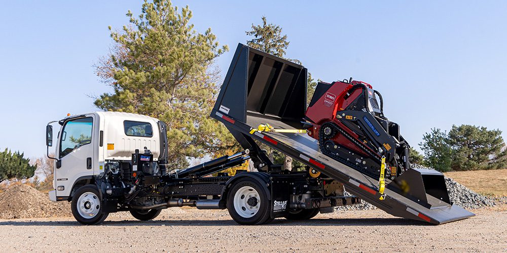 A white cab-over truck with a Stellar Hooklift tilting a flatbed container carrying a red Toro mini skid steer.
