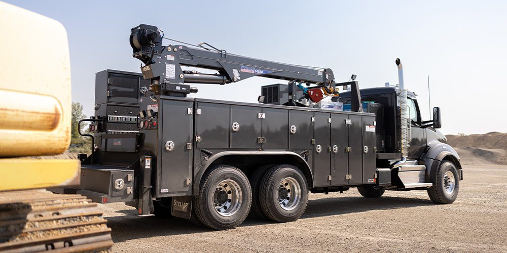 Dark gray tandem-axle Stellar Mechanic Truck with service crane parked on a dusty construction site beside an excavator.