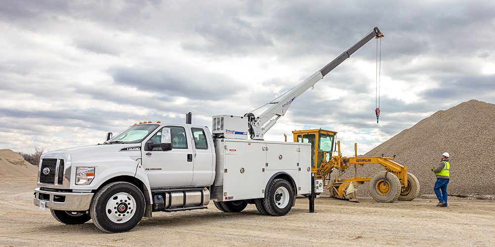 White Stellar mechanic truck with raised telescopic crane operating near a Caterpillar grader on a gravel jobsite.
